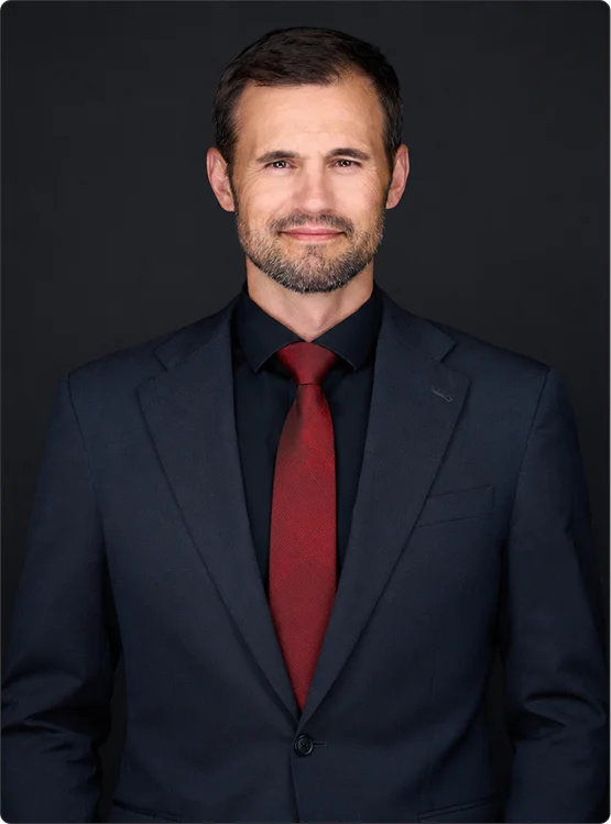 Portrait of a man in a dark suit, black shirt, and red tie against a black background.