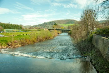 Calm river flowing through rural landscape with hills, bridge, and leafless trees.