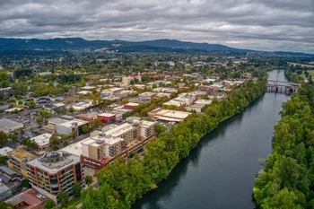 Aerial view of a cityscape with buildings, a river bordered by trees, and distant mountains under a cloudy sky.