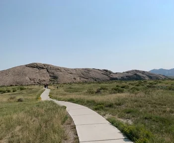 Curved concrete pathway through grassy field leading to large rocky hill under clear blue sky.