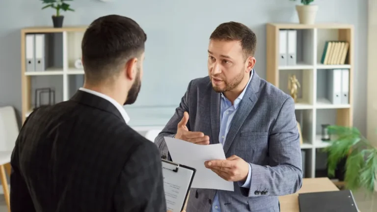 Two professionally dressed men engaged in discussion in an office with shelves and plants.