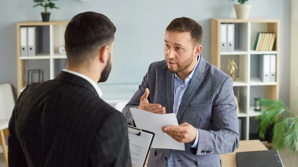 Two professionally dressed men engaged in discussion in an office with shelves and plants.