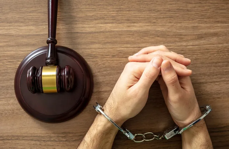 Pair of hands in metal handcuffs beside a wooden judge's gavel on a brown table.