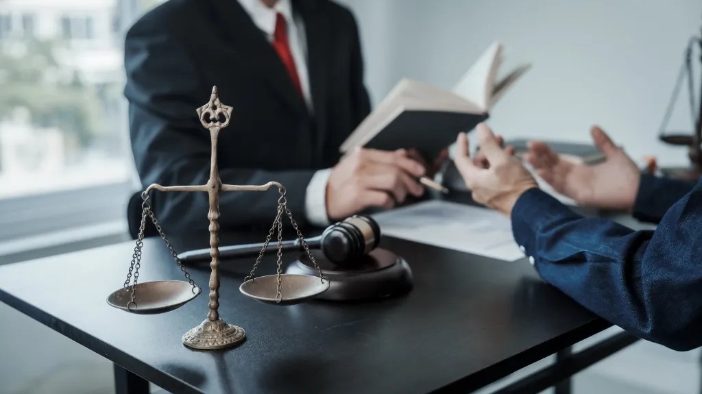 Bronze scales of justice and wooden gavel on desk with two men discussing legal documents.