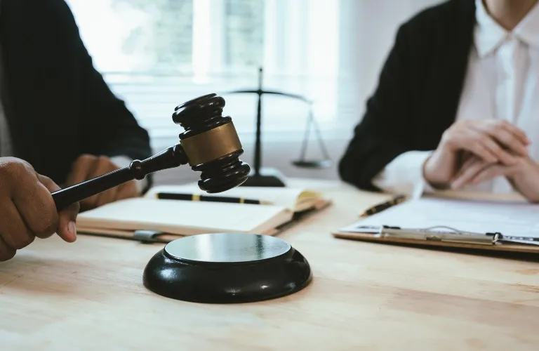 Close-up of a judge's hand holding a wooden gavel above a sound block, with legal documents and scales of justice in the background.