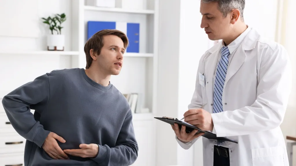 A male patient in a blue sweater holding his abdomen, consulting with a doctor in a white coat.