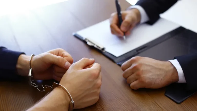 Close-up of a handcuffed individual and a professional writing on a clipboard at a wooden table.