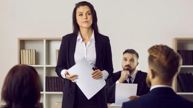 Professional woman presenting in a business meeting with colleagues seated and listening.