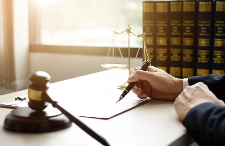 Person in formal attire writing on paper at desk with gavel, scales of justice, and law books.