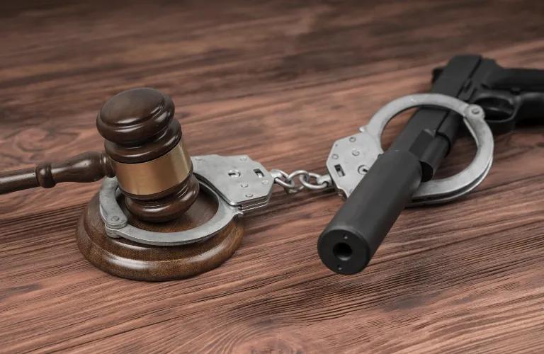 Wooden judge's gavel and black handgun connected by silver handcuffs on wooden surface.