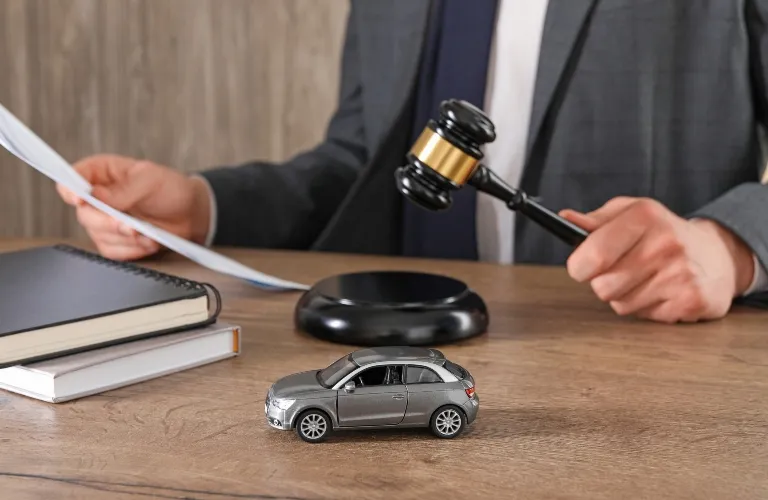 Person in gray suit holding judge's gavel with toy car and books on wooden desk.