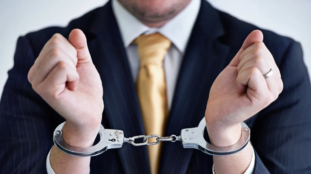 Man in dark pinstripe suit and gold tie with hands cuffed in silver handcuffs.