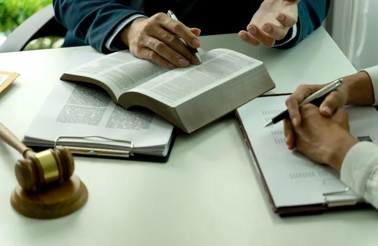 Two individuals reviewing legal documents, one holding a pen over an open book, with a wooden gavel on the table.