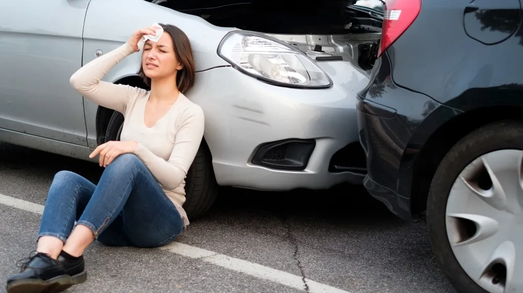 Woman sitting on the ground beside two cars involved in a rear-end collision, holding her forehead.