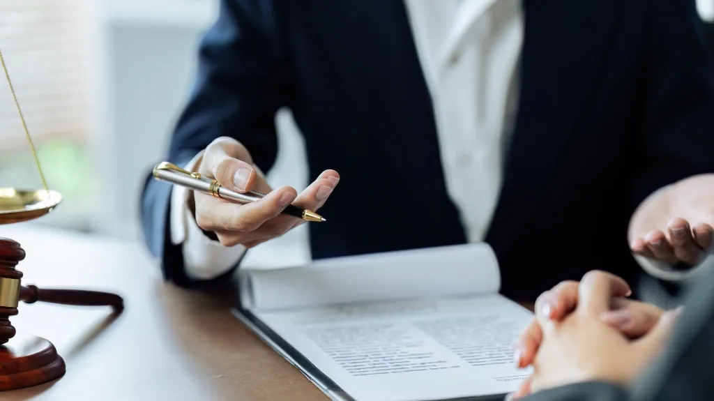 Professional in dark suit explaining document details with pen, legal scales and gavel on table.