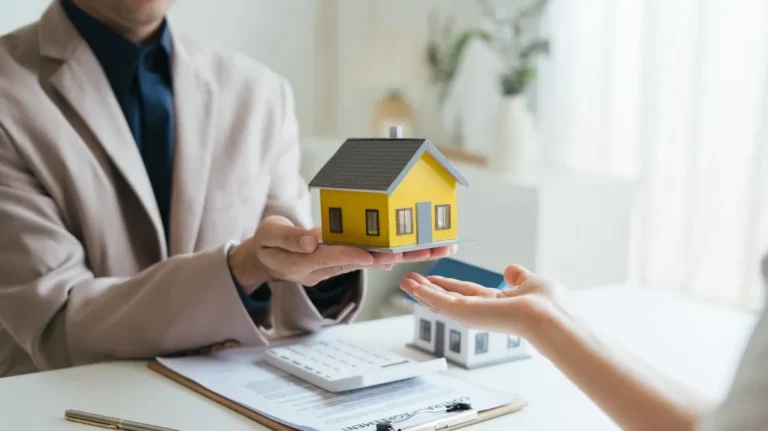 Real estate agent in beige jacket presenting yellow house model to client over contract and calculator.