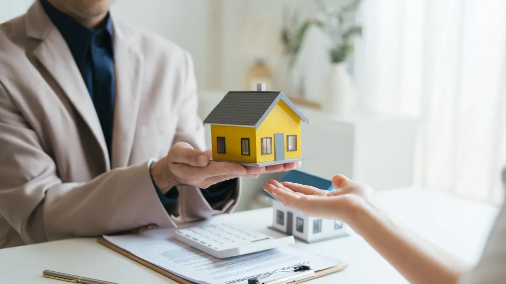 Real estate agent in beige jacket presenting yellow house model to client over contract and calculator.