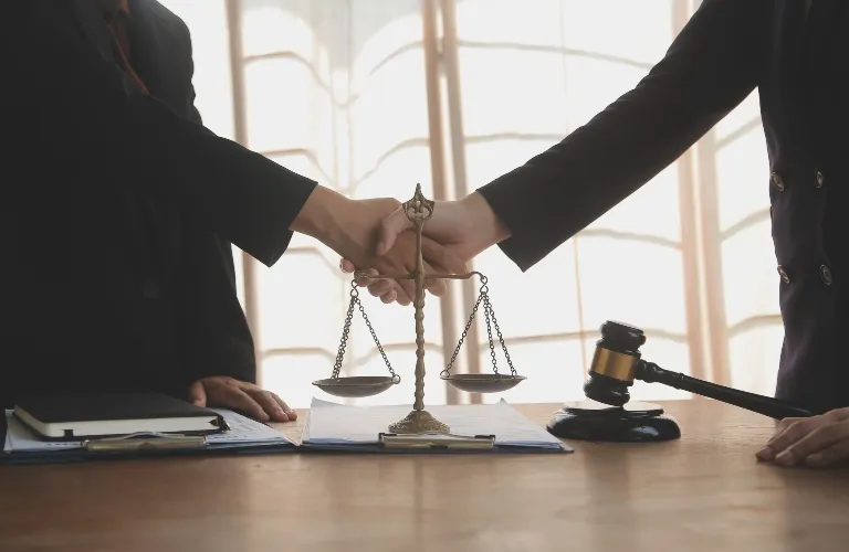 Two individuals in formal attire shaking hands over a desk with legal scales and a judge's gavel.
